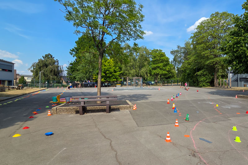 Großer Schulhof mit Bänken, einem Baum in der Mitte und bunten Hütchen, die auf dem Asphalt verteilt sind.