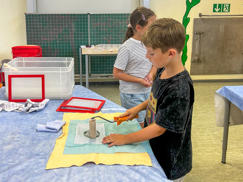 Ein Junge mit schwarzem T-Shirt bearbeitet ein quadratisches Stück Stoff mit einem Werkzeug auf einem Tisch, im Hintergrund ist ein Mädchen mit gestreiftem T-Shirt und eine grüne Tafel zu sehen.