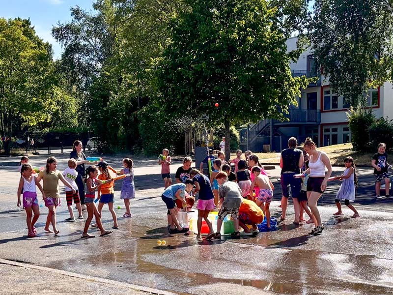 Gruppe von Kindern und Erwachsenen spielt mit Wasser und Eimern auf einem sonnigen Schulhof mit Bäumen und Schulgebäude im Hintergrund.