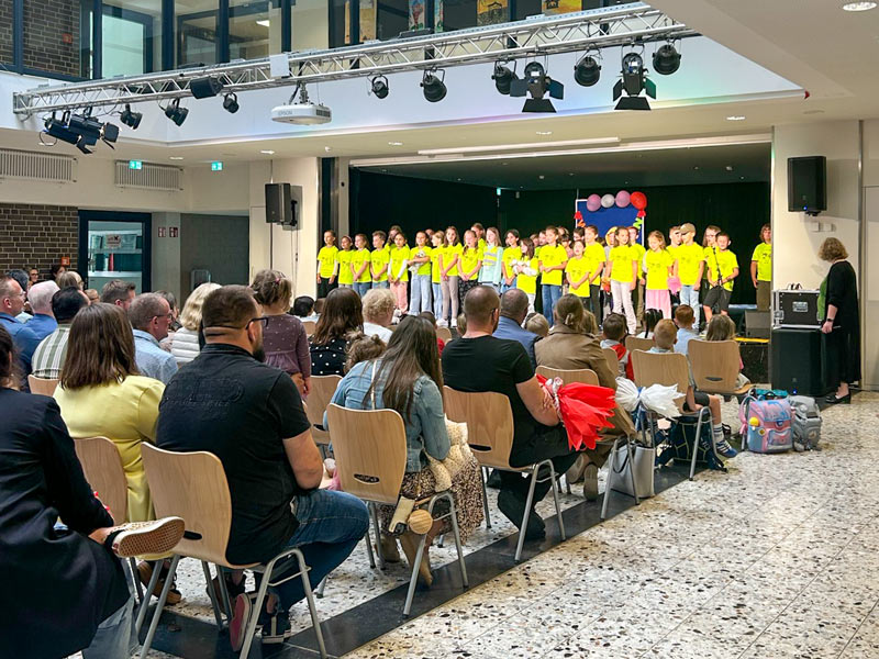 Kinder in gelben T-Shirts stehen auf einer Bühne und singen vor einem sitzenden Publikum in einem hellen Raum.