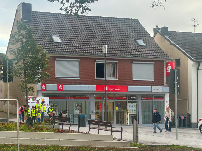 Zweigeschossiges Backsteingebäude mit Sparkassenfiliale im Erdgeschoss und mehreren Personen auf dem Gehweg davor.